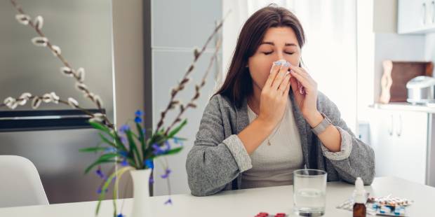 Spring allergy. Young woman sneezing because of flowers surrounded with pills on kitchen. Seasonal allergy concept.